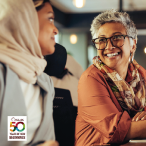Two women share eye contact and big smiles. One has short grey hair and glasses and the other wears a hijab.