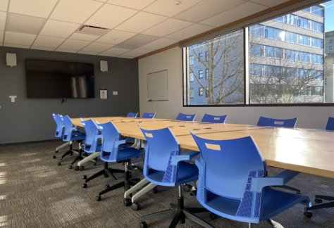 A table, 12 chairs, a monitor on the wall, and a large window inside the Ash Street Room at New Westminster Public Library's Main Branch.