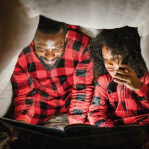 A parent and child read a book with a flashlight in a blanket fort.