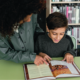 A child reads a book sitting at a table while an adult helps by pointing to some text.