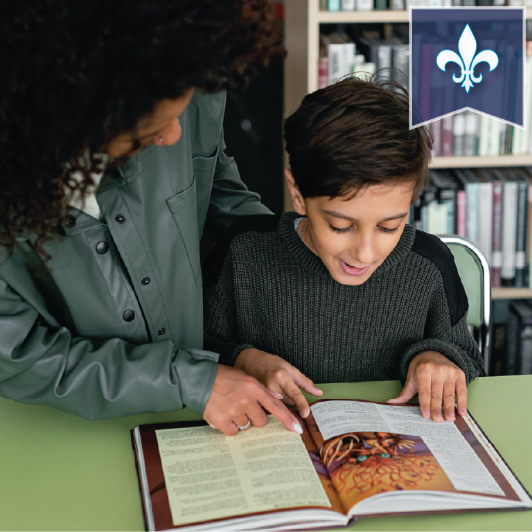 A child reads a book sitting at a table while an adult helps by pointing to some text.