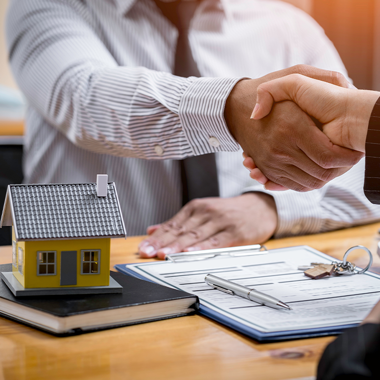 two people shake hands across a desk with signed papers, a set of keys and a tiny toy house.