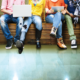 Four young people sit on a bench, with only their lower bodies visible. They are sharing electronic devices and wearing youthful clothing.