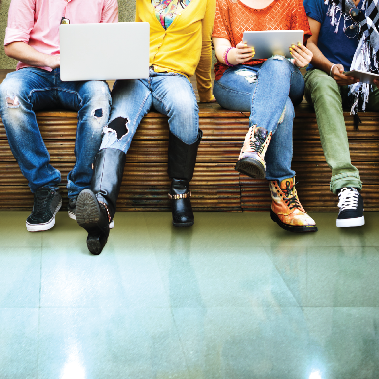 Four young people sit on a bench, with only their lower bodies visible. They are sharing electronic devices and wearing youthful clothing.