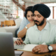 A couple sits in front of a computer doing paperwork. The husband wears a black turban and is entering information on the computer and the wife is slightly behind him leaning on his shoulder.