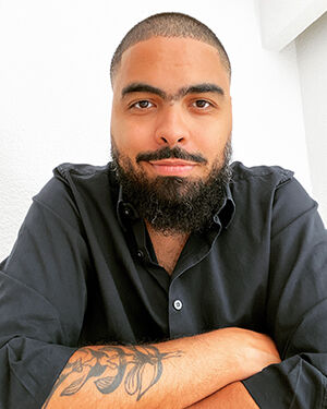 A light skinned black man with very short hair and wearing a black shirt looks directly into the camera.