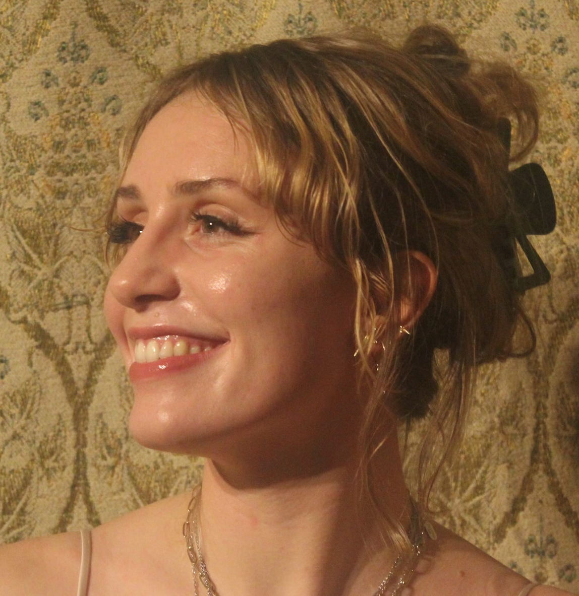 with a gold background, a smiling woman with blonde hair looks toward the left. She has gold chains around her neck and bare shoulders.