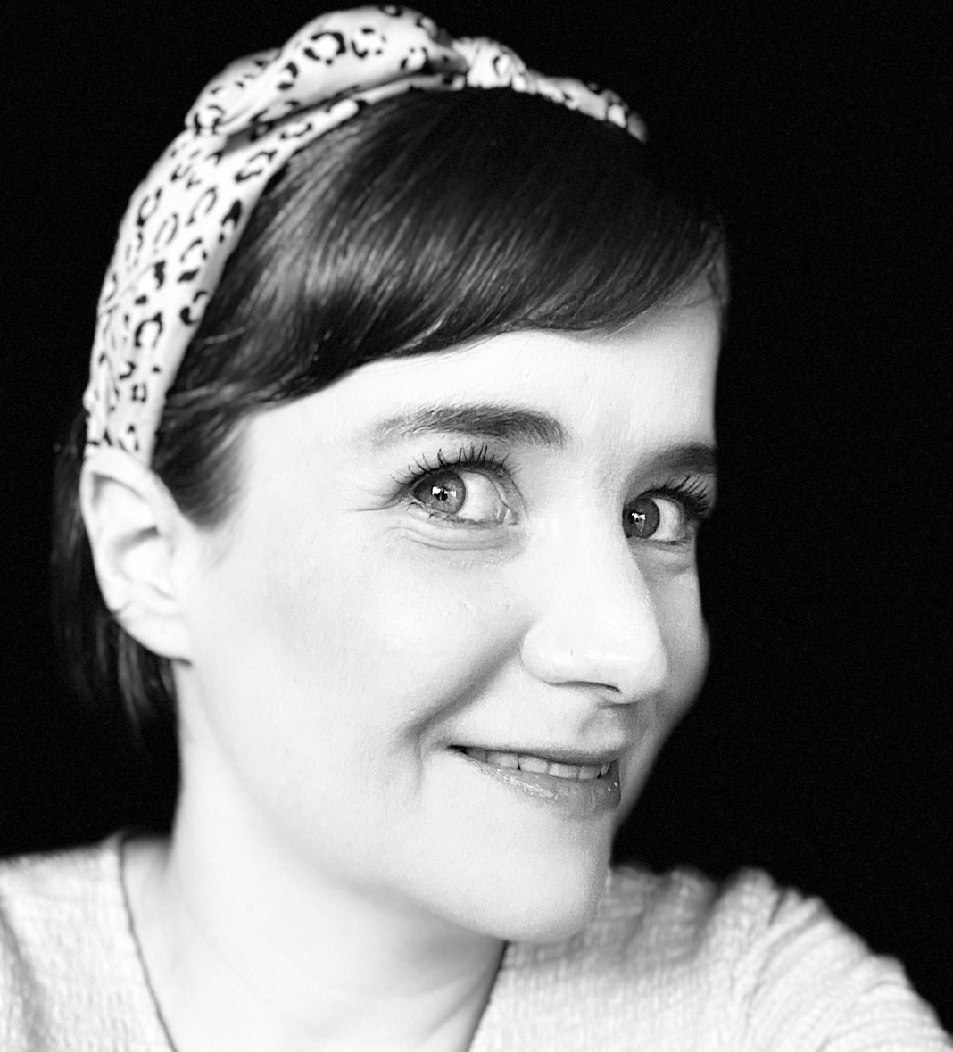 In a black and white photo, a woman with a thick headband and dark hair looks sideways into the camera with a slight smile.
