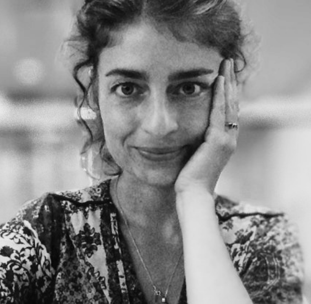 In a black and white photograph, a woman with dark curly hair gazes into the camera, her chin resting on her hand.