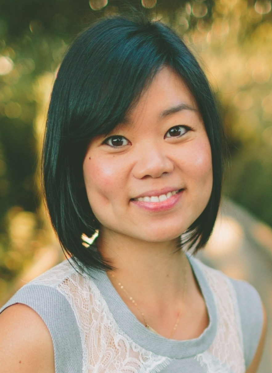 Against a natural backdrop, a dimpled Asian woman in a light blue dress smiles at the camera.