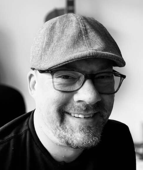A black & white photo of a man with a short beard and wearing a cap and glasses, smiling and looking into the camera. Some guitars hang on the wall behind him.
