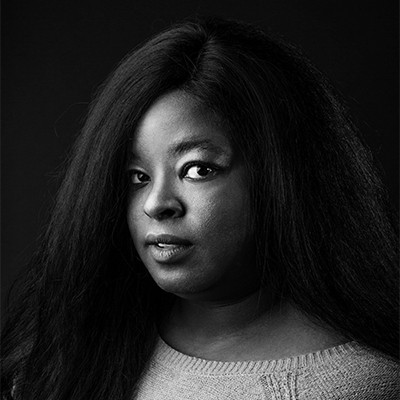 Against a black background, a black woman with long hair looks sideways into the camera.