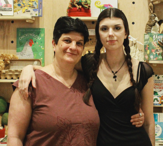 A mother and daughter, both with dark hair, embrace each other and look toward the camera, in front of a book store display.