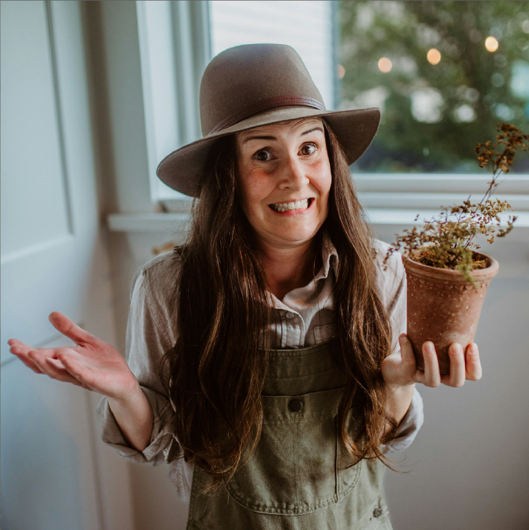 A white woman with long brown hair and a perplexed expression holds up a dead plant in a pot and lifts her other hand in disbelief.