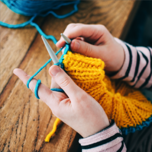 A pair of hands is knitting a project with a wide yellow band, starting on a bright turquoise section. The yarn is wound around the person's fingers and the knitting needles are visible.