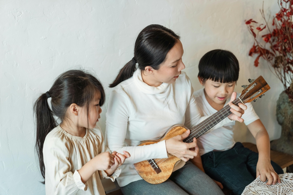 Mother plays a ukulele while two children observe and listen.