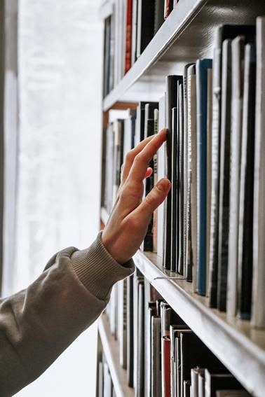 A left hand reaches for a book on a shelf. 