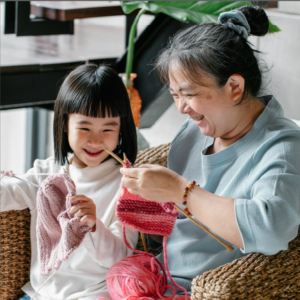 A young girl and a grandmother are knitting together and have big smiles.