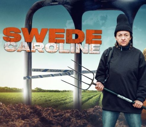 A fierce looking woman with a pitchfork and wearing a toque stands in front of giant fork tines.