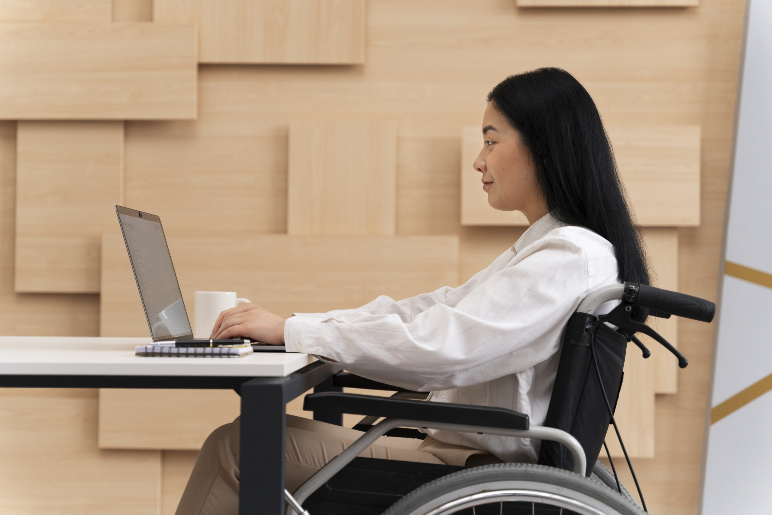 Young adult sitting in a wheelchair works on a laptop computer with a modern wooden wall in the background.