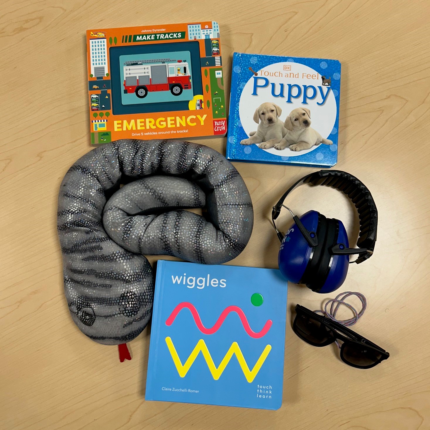 Three tactile board books, a weighted snake, headphones, and sunglasses on a wooden table.