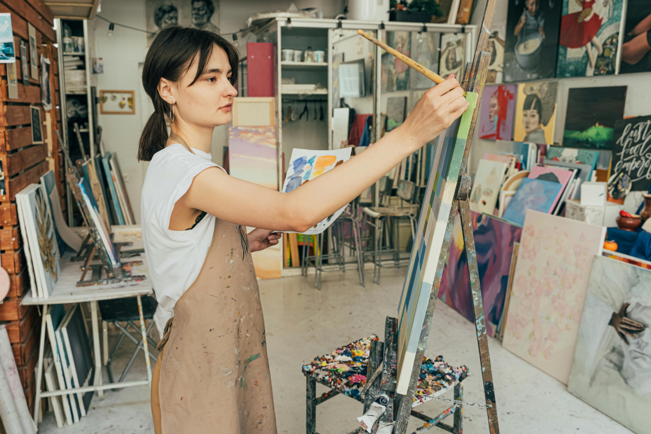 A teen paints on a medium-sized canvas in a studio filled with art supplies and other artworks.