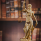 Closeup of a "Lady Justice" figurine, who wears a white toga with brass-coloured trim and a brass-coloured blindfold. She is holding scales in her right hand and a sword in her left hand.