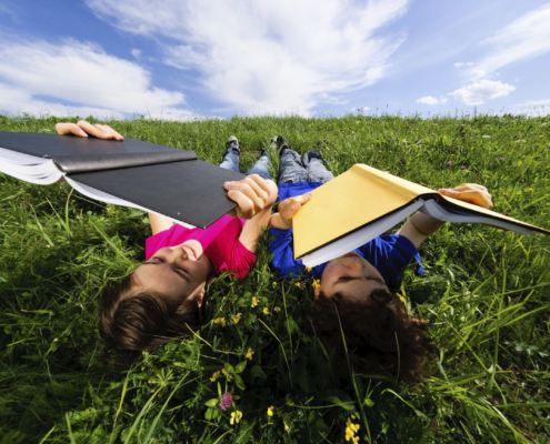 Two children lie on their backs in a grassy field, reading books held above their faces.